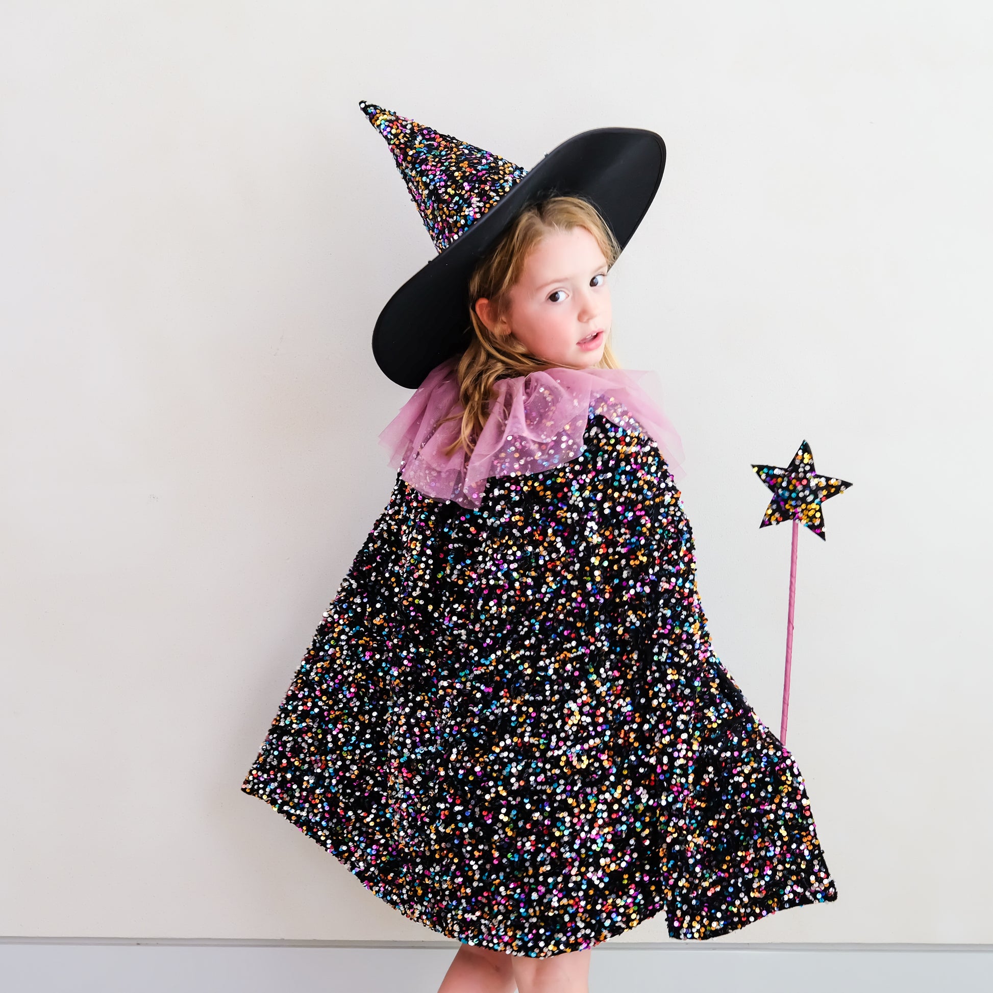 A young girl wearing a black witch costume cape covered in multicolor glitter, a black pointed witch hat, and a pink ruffled collar, holding a matching glittery star wand against a plain white wall.
