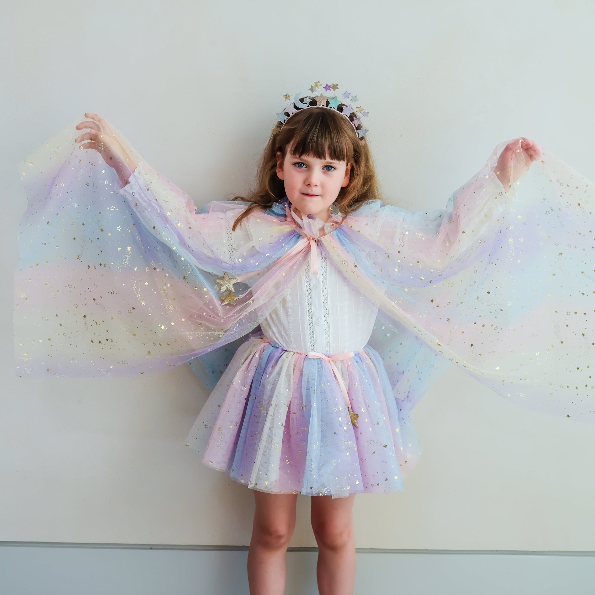 A young girl wearing a pastel unicorn-themed costume dress with a matching sheer cape, tiara, and sparkly star details, posing with her arms outstretched against a plain light gray wall.