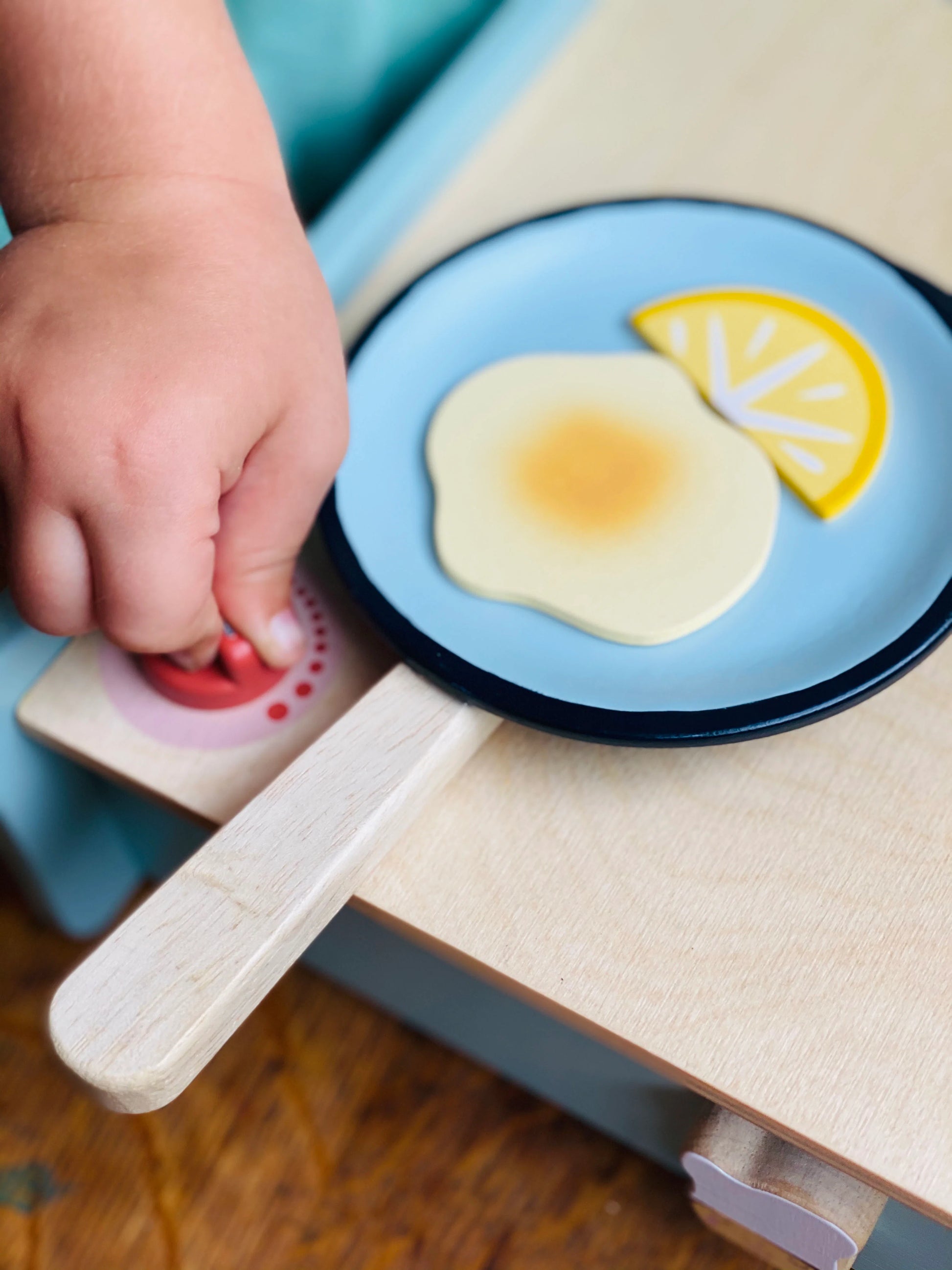 Child's hand turning knob on wooden toys stove whilst a wooden pancake and lemon slice sit in frying pan