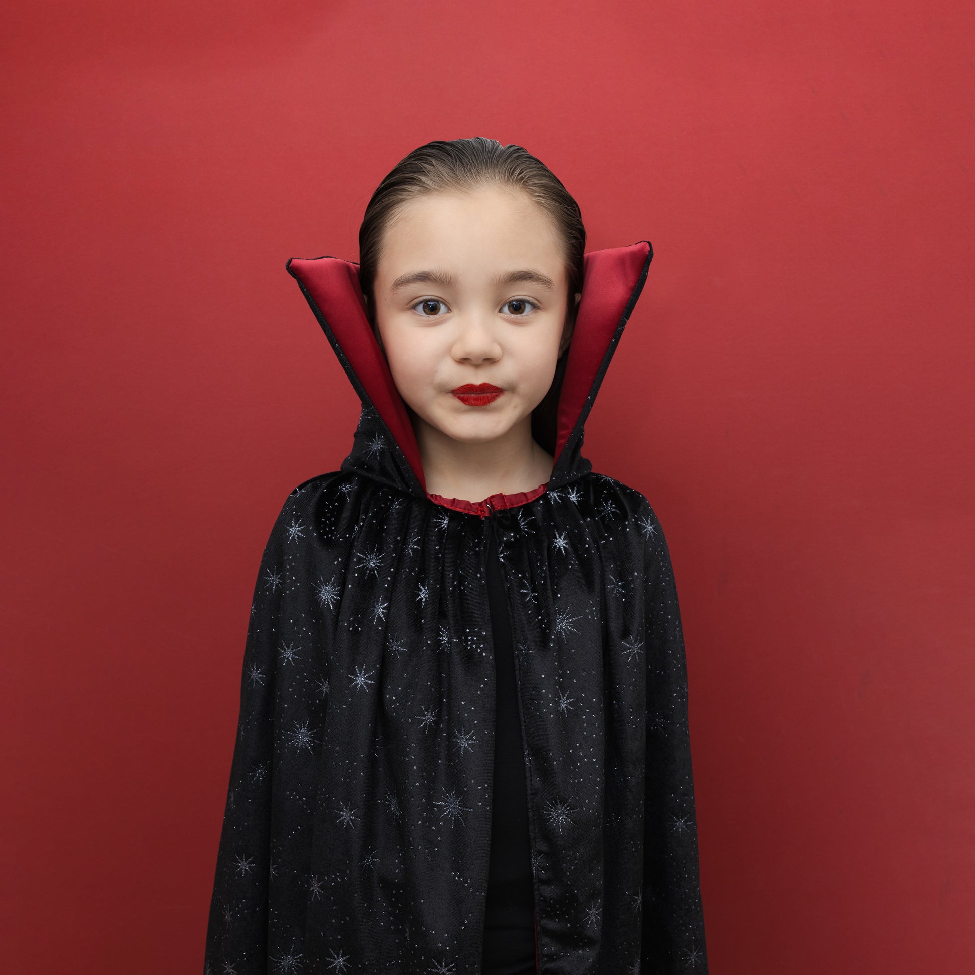 A young girl wearing a black vampire cape with silver star patterns and a red inner collar, posing against a solid red background.