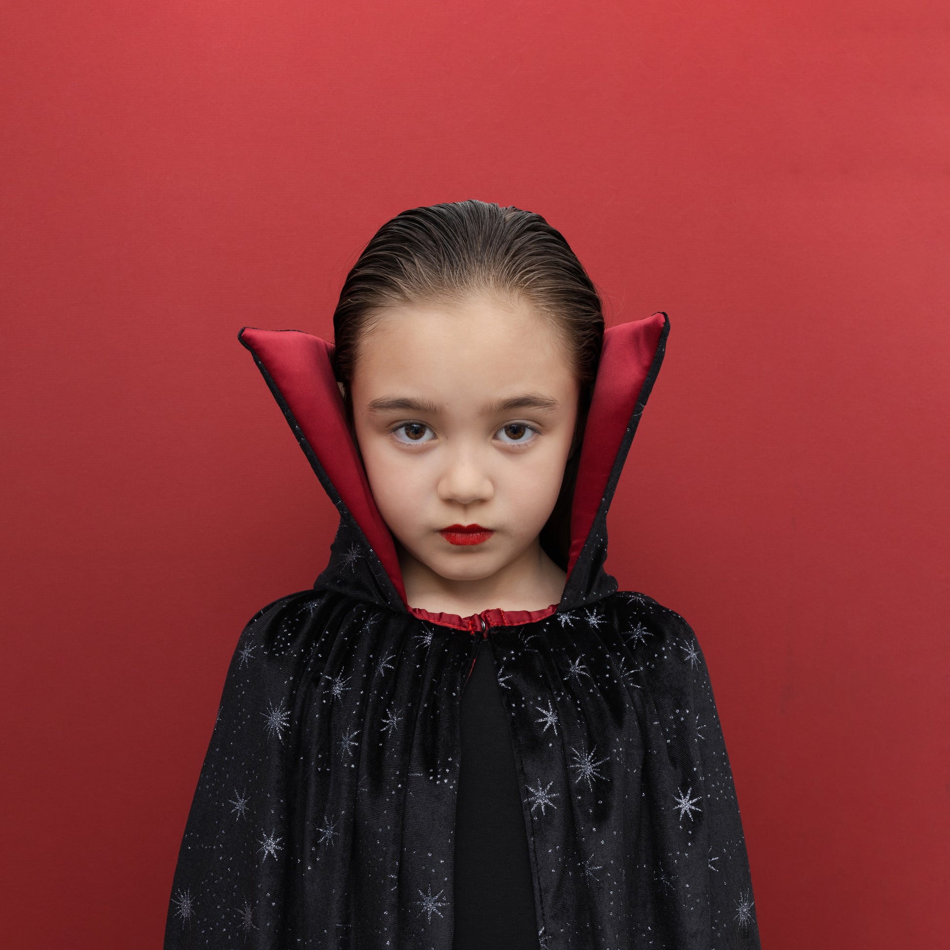 A young girl with slicked-back dark hair wearing a black vampire cape with silver star patterns and a red inner collar, posing against a solid red background.