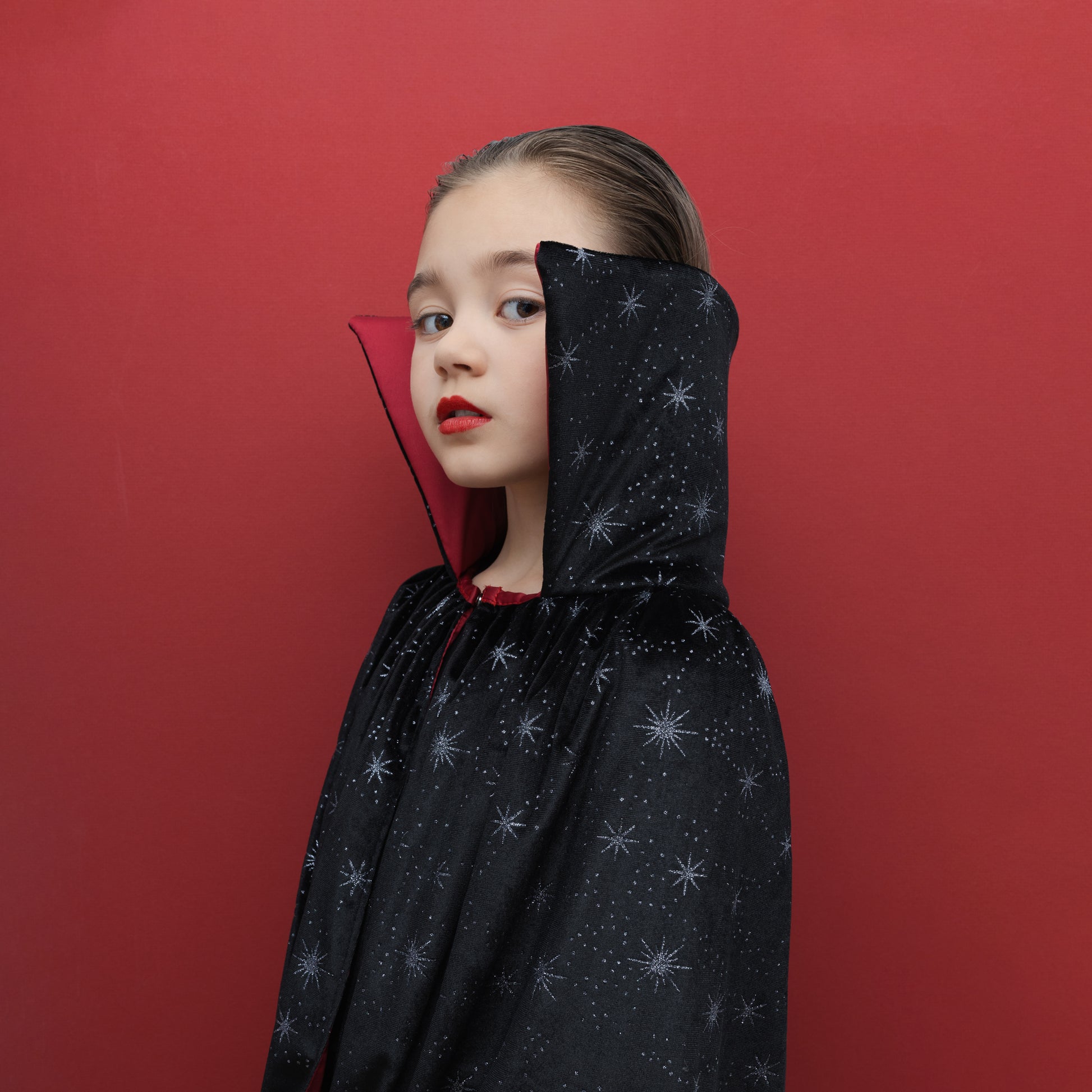 A young girl wearing a black hooded cape with a silver star pattern, posed against a solid red background.