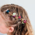 A close-up of a child's braided brown hair styled with several colorful hair accessories, including a blue scrunchie, a wooden snail clip, a purple scrunchie, a red ladybug clip, and a burgundy flower clip, against a plain light gray background.