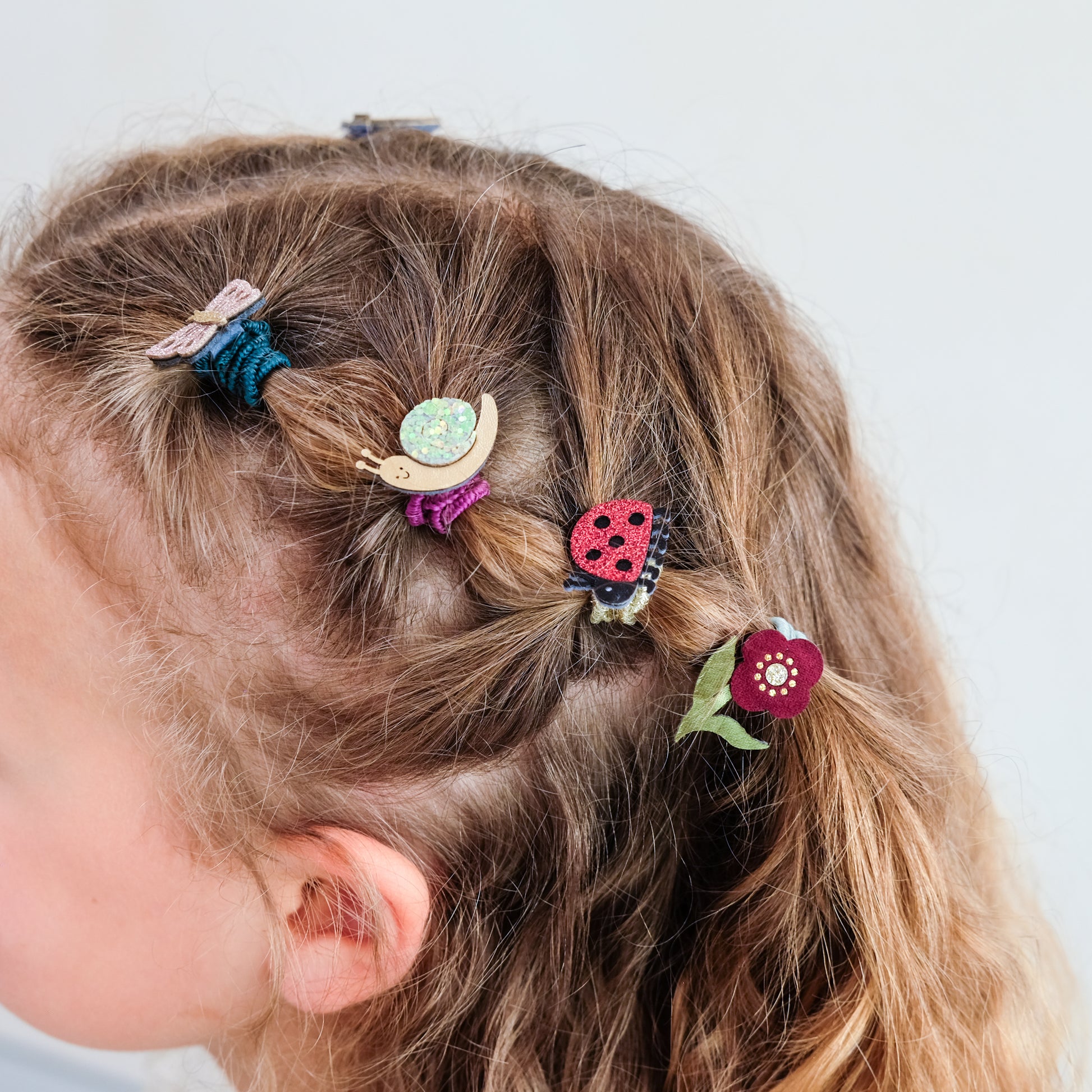 A close-up of a child's braided brown hair styled with several colorful hair accessories, including a blue scrunchie, a wooden snail clip, a purple scrunchie, a red ladybug clip, and a burgundy flower clip, against a plain light gray background.