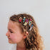 A side-profile portrait of a young girl with curly light brown hair wearing five colorful decorative hair clips shaped like a ladybug, striped shoes, a teapot, a flower, and a butterfly against a plain light background.