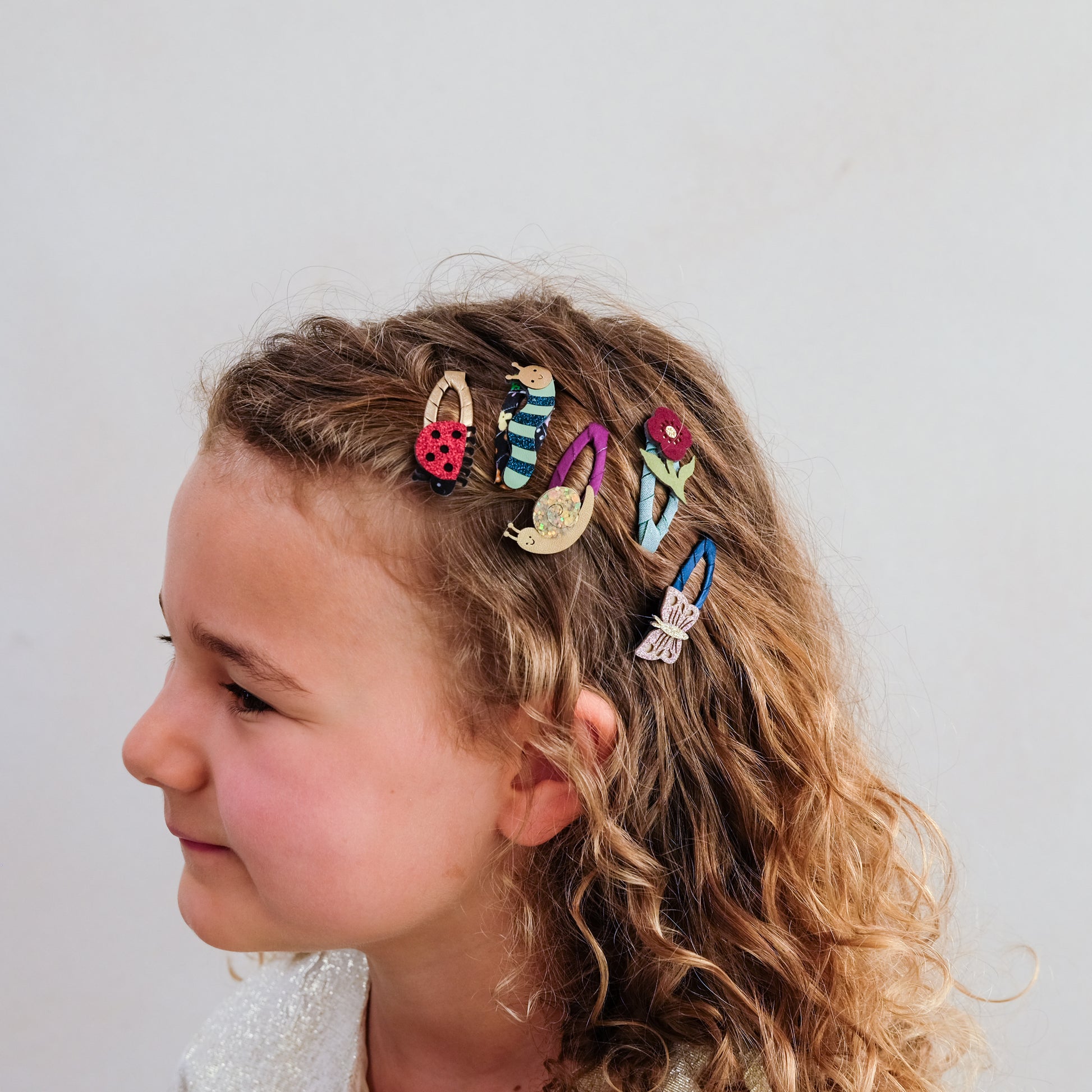 A side-profile portrait of a young girl with curly light brown hair wearing five colorful decorative hair clips shaped like a ladybug, striped shoes, a teapot, a flower, and a butterfly against a plain light background.