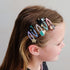 A side-profile view of a young girl with brown hair wearing a set of five colorful, whimsical hair clips. The clips feature cute cartoon designs including a black cat with a bow, a striped character, a pink witch, a blue moon, and a gold glitter star, all attached to clear plastic claw clips.