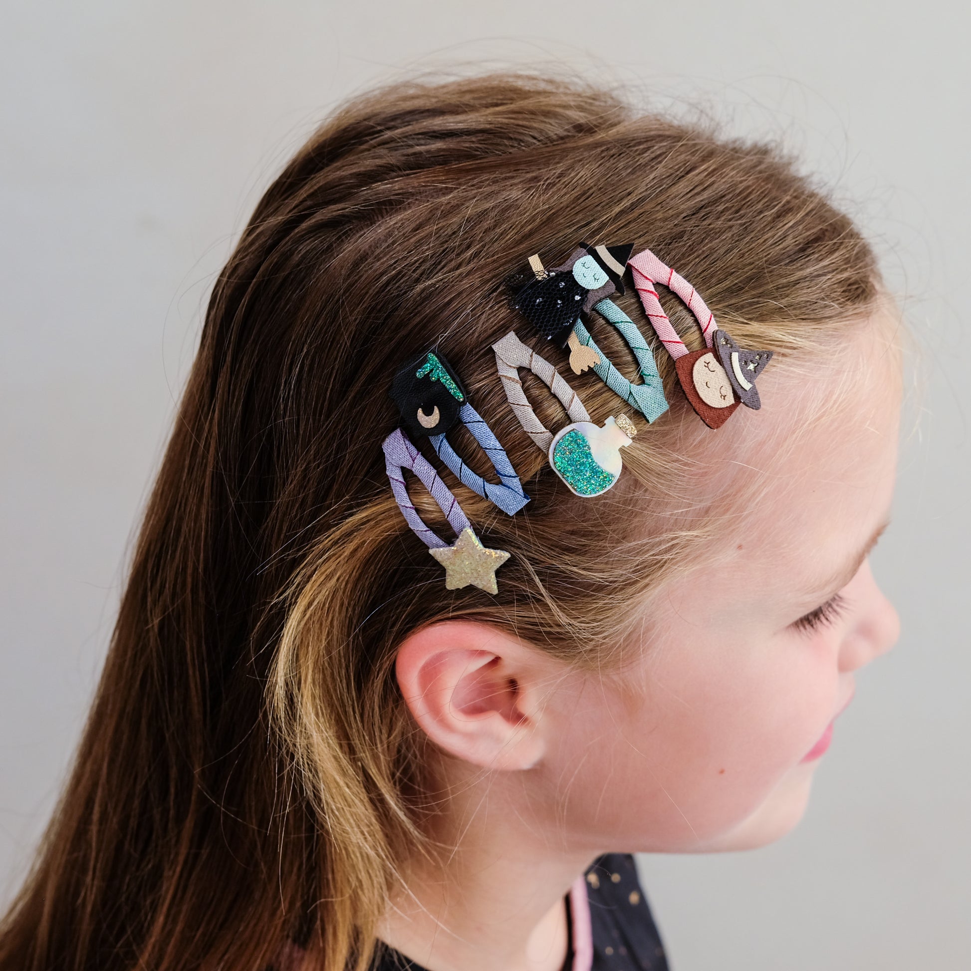 A side-profile view of a young girl with brown hair wearing a set of five colorful, whimsical hair clips. The clips feature cute cartoon designs including a black cat with a bow, a striped character, a pink witch, a blue moon, and a gold glitter star, all attached to clear plastic claw clips.
