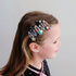 A side-profile view of a young girl with brown hair wearing a set of colorful, whimsical hair clips. The clips feature cute character designs including a blue bird, a green frog, a pink owl, and a gold star, attached to elastic bands.