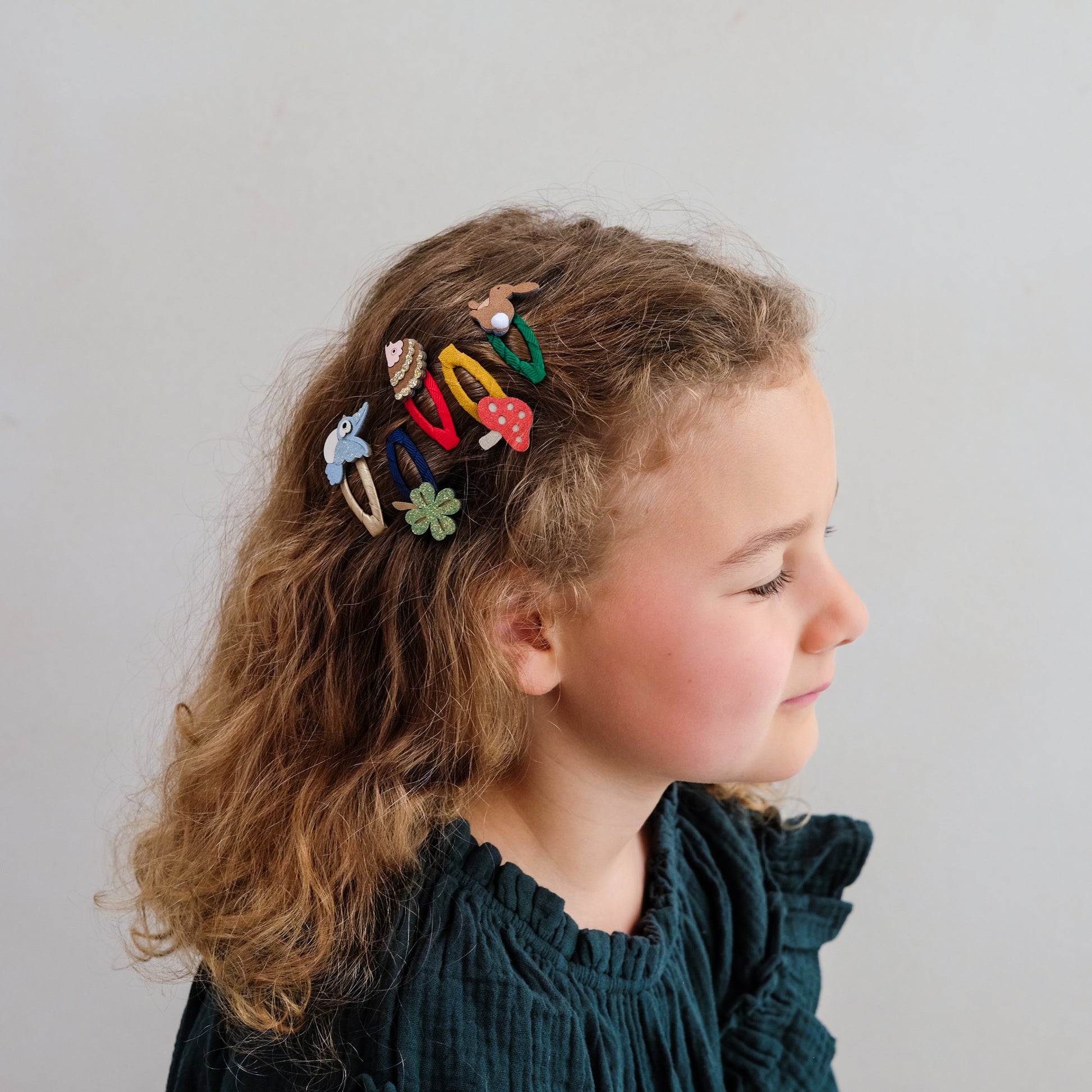 A young girl with curly light brown hair is shown in profile wearing a set of colorful mushroom-themed hair clips. The clips feature whimsical designs including a blue mushroom, a red mushroom, a green flower, and others in brown, yellow, and white, attached to a black hair claw clip.