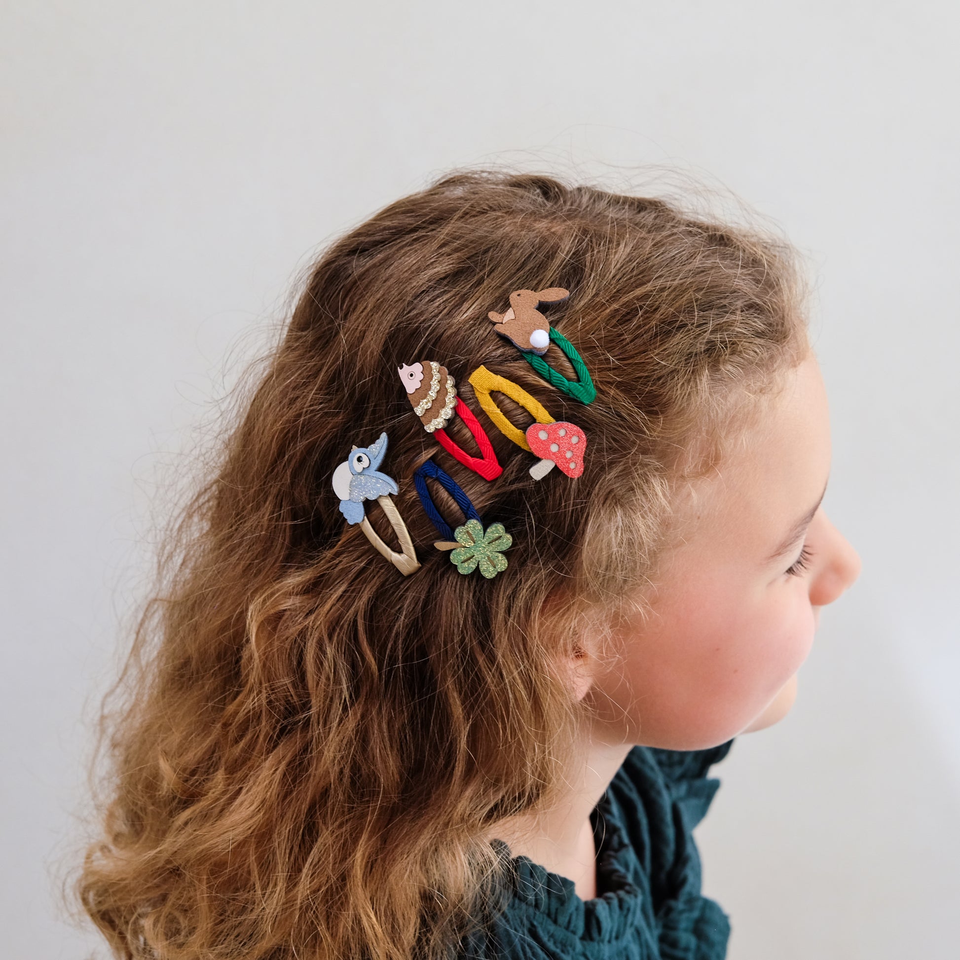 A side-profile view of a young girl with wavy light brown hair wearing several colorful mushroom-themed hair clips against a plain light gray background.