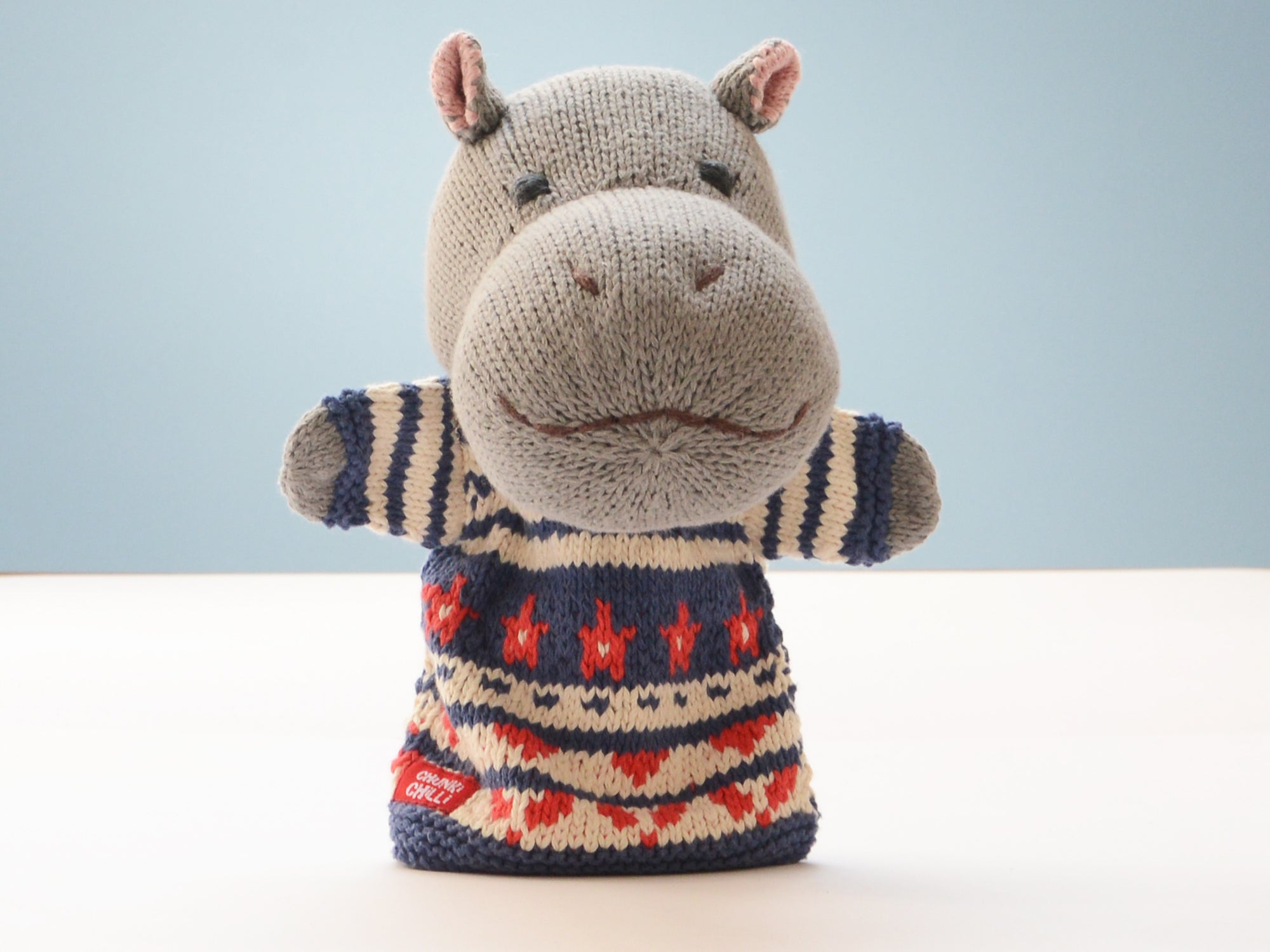 A knitted hippo-shaped hand puppet wearing a patterned sweater, standing on a white surface against a light blue background.
