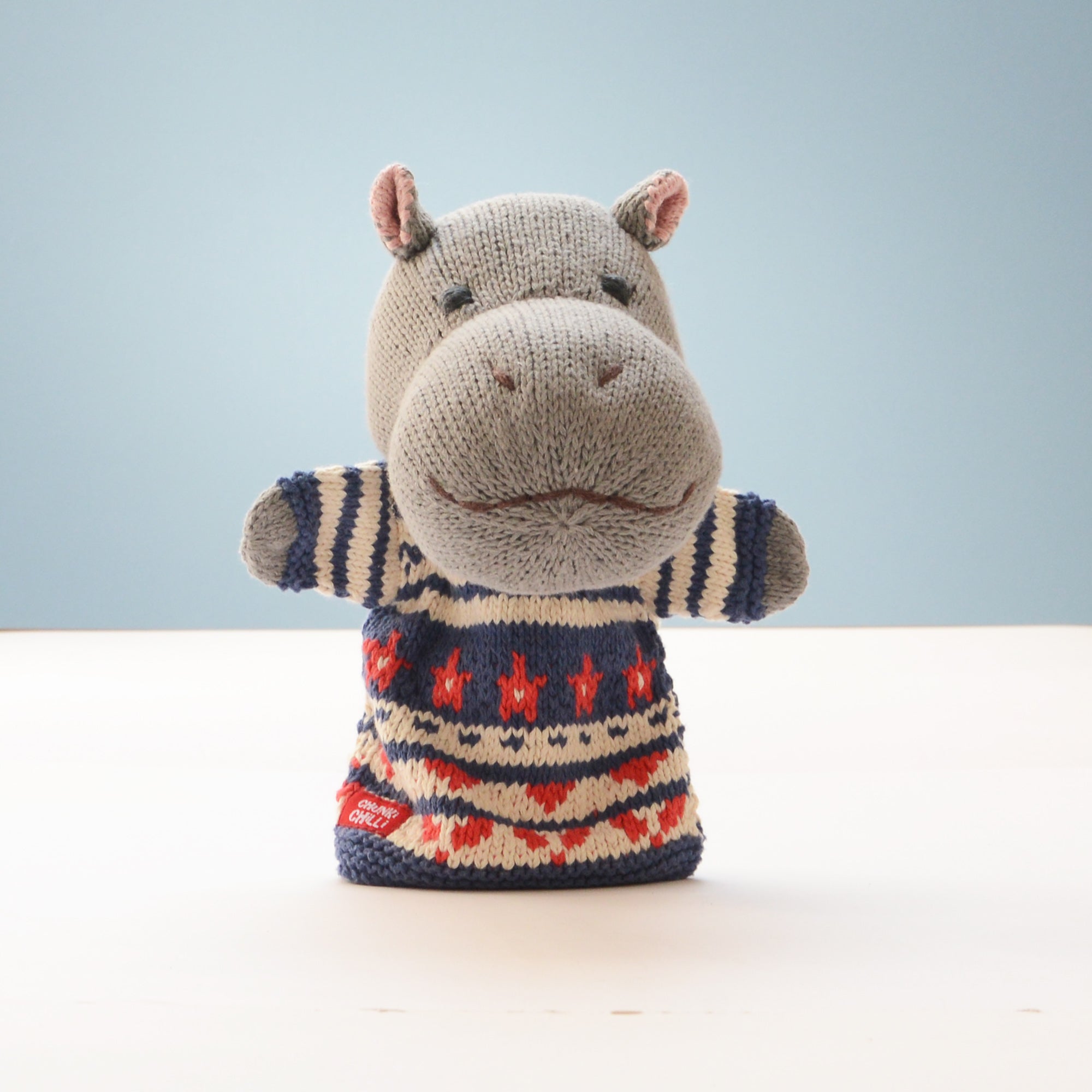 A knitted hippo-shaped hand puppet wearing a patterned sweater, standing on a white surface against a light blue background.