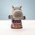 A knitted hippo-shaped hand puppet wearing a patterned sweater, standing on a white surface against a light blue background.