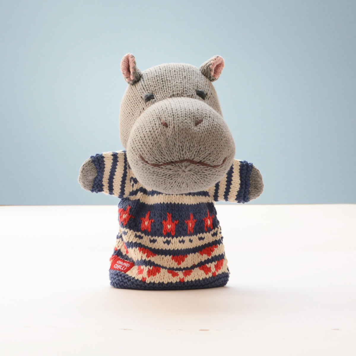 A knitted hippo-shaped hand puppet wearing a patterned sweater, standing on a white surface against a light blue background.