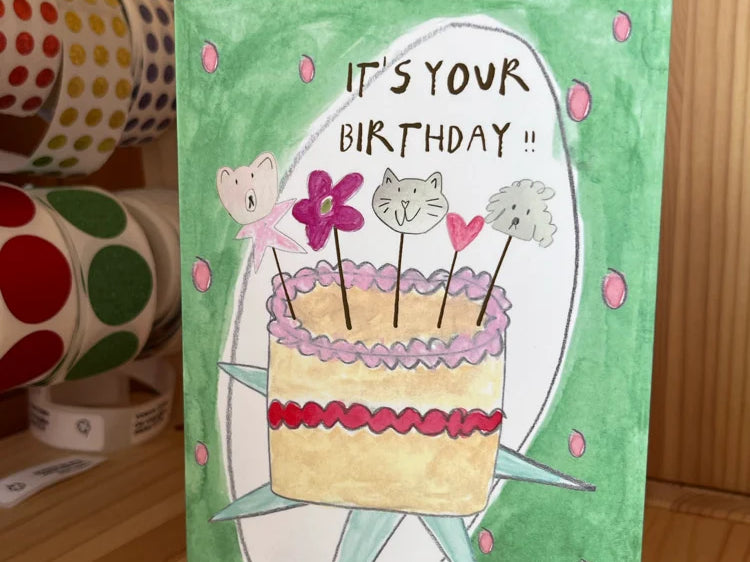 A handmade watercolor birthday card with a green background, featuring a drawing of a cake with cat faces on top and the text 'IT'S YOUR BIRTHDAY !!'. The card is standing on a wooden surface with washi tape rolls and stickers in the background.