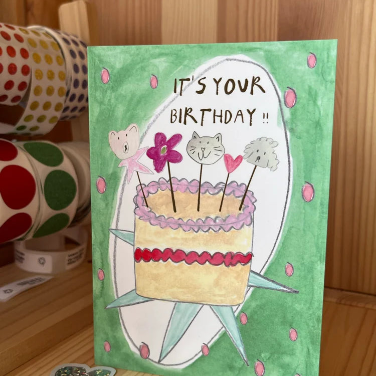 A handmade watercolor birthday card with a green background, featuring a drawing of a cake with cat faces on top and the text 'IT'S YOUR BIRTHDAY !!'. The card is standing on a wooden surface with washi tape rolls and stickers in the background.