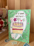 A handmade watercolor birthday card with a green background, featuring a drawing of a cake with cat faces on top and the text 'IT'S YOUR BIRTHDAY !!'. The card is standing on a wooden surface with washi tape rolls and stickers in the background.