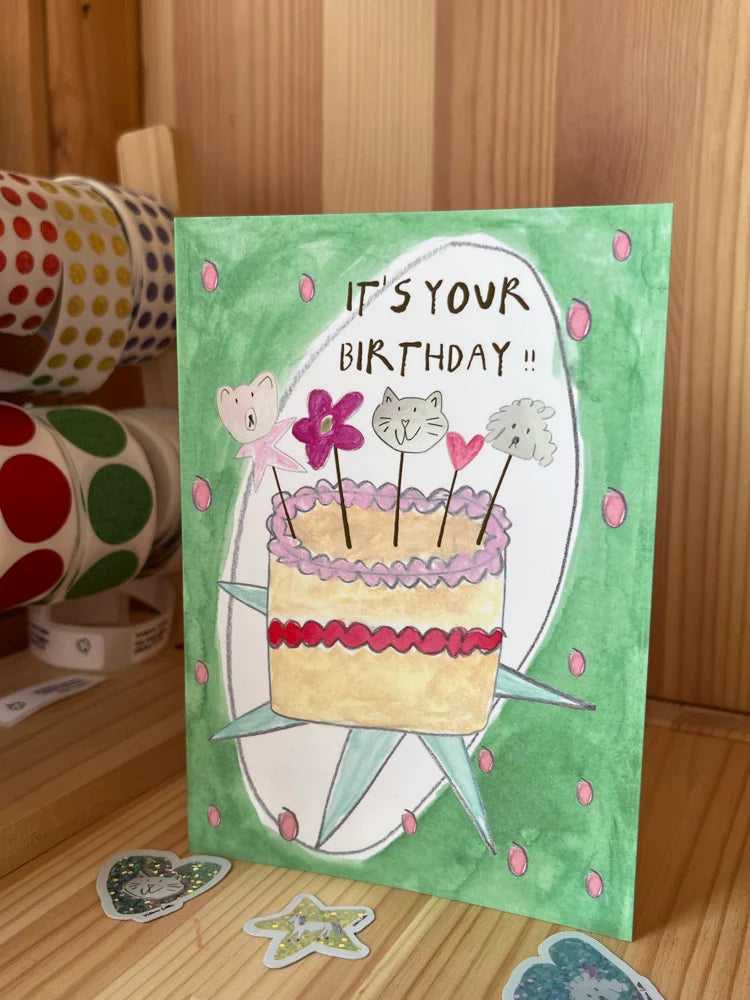A handmade watercolor birthday card with a green background, featuring a drawing of a cake with cat faces on top and the text 'IT'S YOUR BIRTHDAY !!'. The card is standing on a wooden surface with washi tape rolls and stickers in the background.